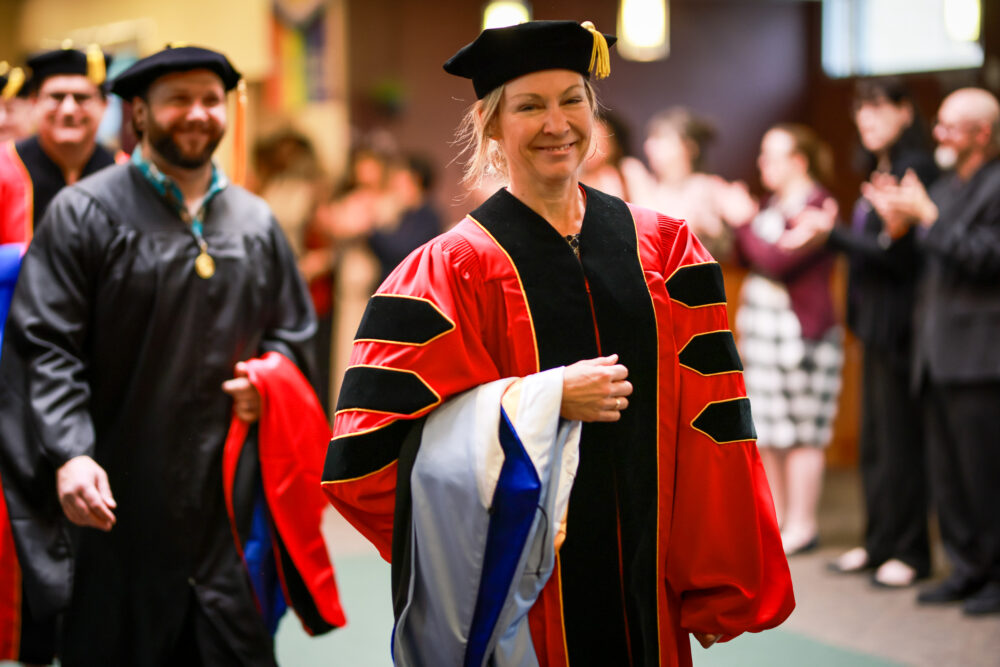 Smiling woman wearing academic regalia walks with her colleagues to receive her PhD